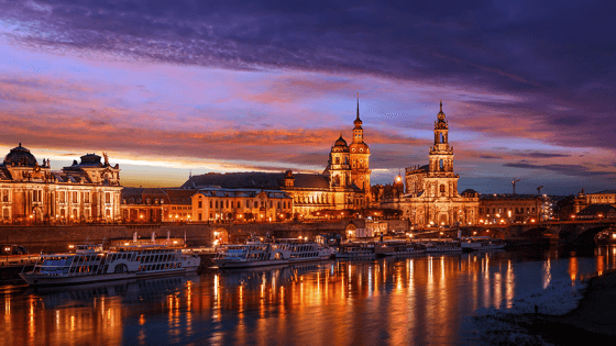 Man sieht hell erleuchtete Gebäude. Es ist Sonnenuntergang. Vor den Gebäuden sieht man einen Fluss