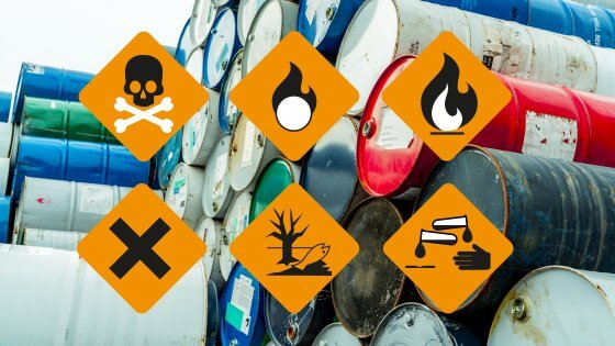 Auf dem Bild sieht man im Vordergrund einige der Piktogramme für Gefahrstoffe. Im Hintergrund sieht man gestapelte Metalltonnen.